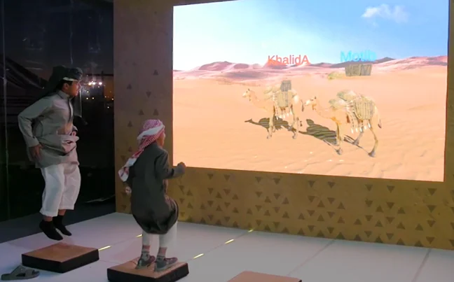 Two visitors use raised pads in front of a large camel-race display.